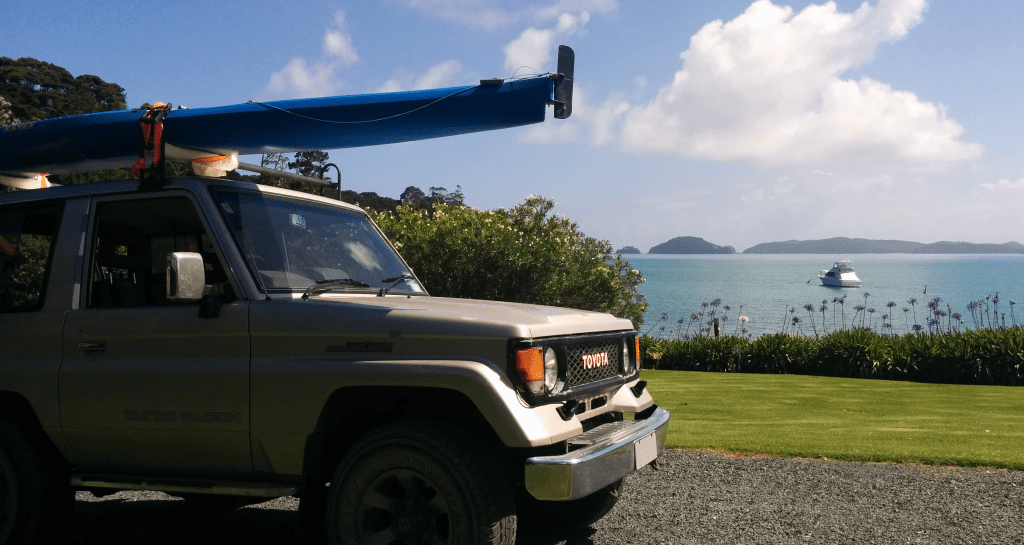 A land cruiser with a kayak on top in front of a sunny beach with a boat
