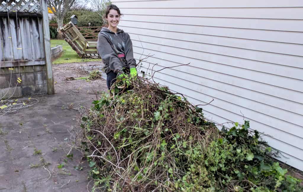 brunette woman in brown sweater dragging weeds across a paved backyard